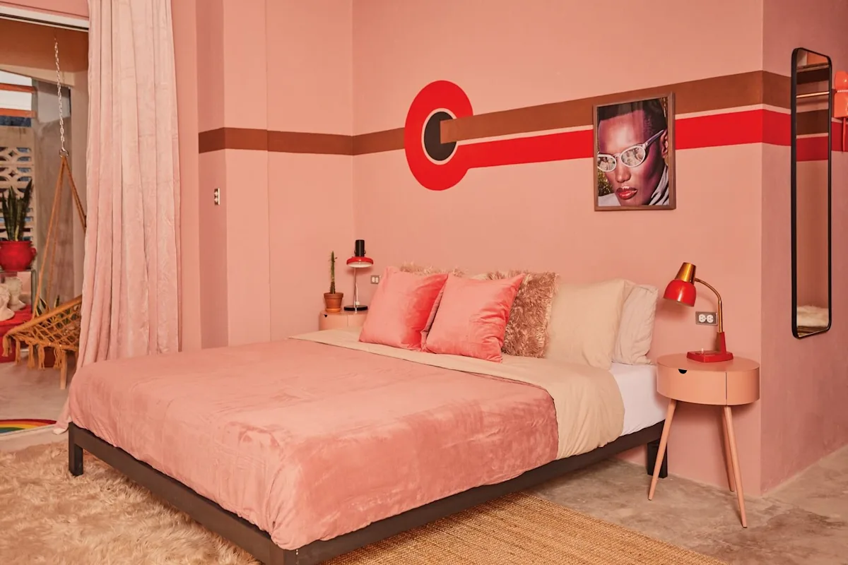 A retro-themed bedroom with pink walls, a pink bedspread, a red accent stripe, and a framed portrait above the bed.