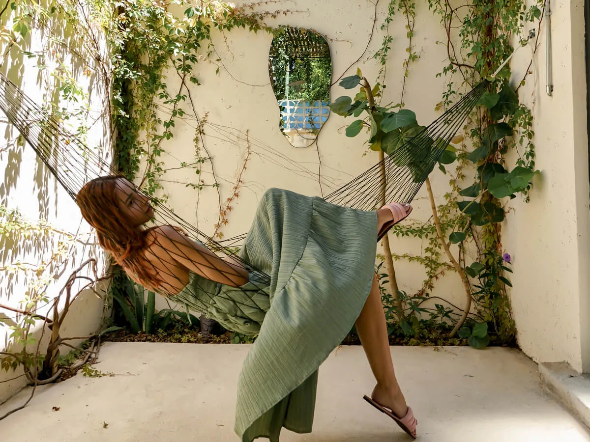 A woman in a sage green dress relaxing in a black rope hammock in a sunlit courtyard framed by climbing ivy vines and a wall-mounted mirror.