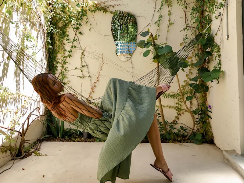 A woman in a sage green dress relaxing in a black rope hammock in a sunlit courtyard framed by climbing ivy vines and a wall-mounted mirror.