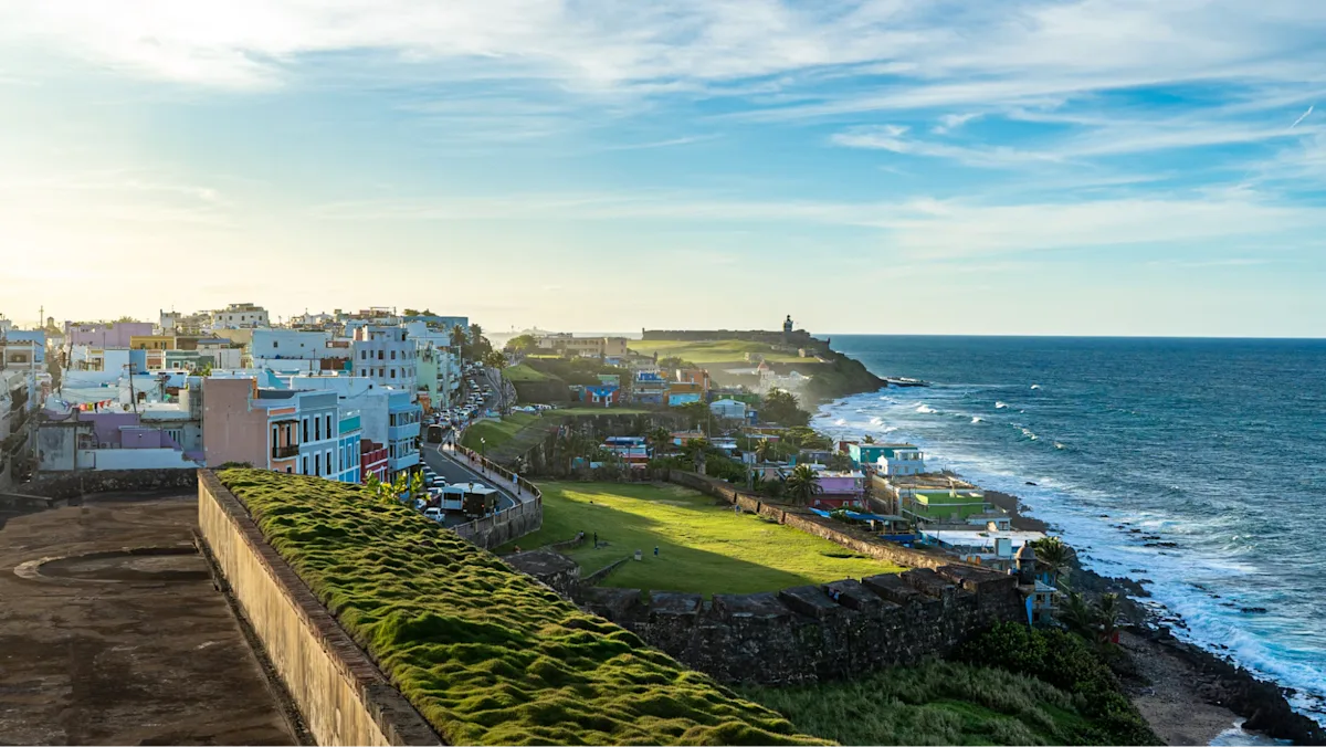 A coastal view of Old San Juan, Puerto Rico, with colorful buildings, historic stone fort walls, and the ocean stretching along the shoreline under a bright blue sky.
