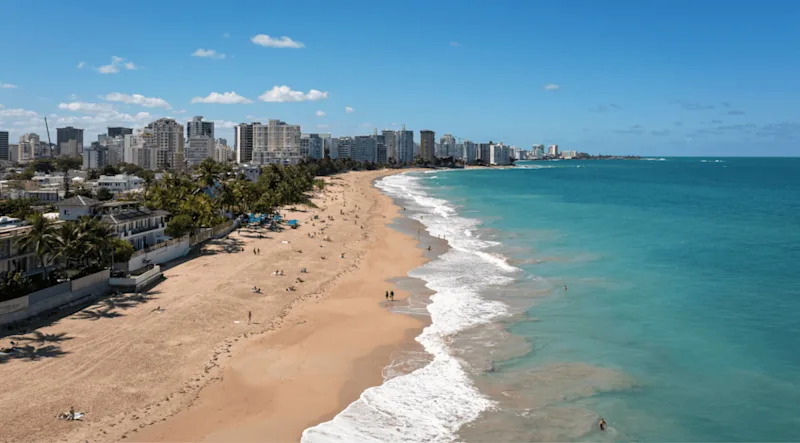 A San Juan beach seen from the air, with the high-rise hotels and apartment towers of the city's beach strip running behind the sand.