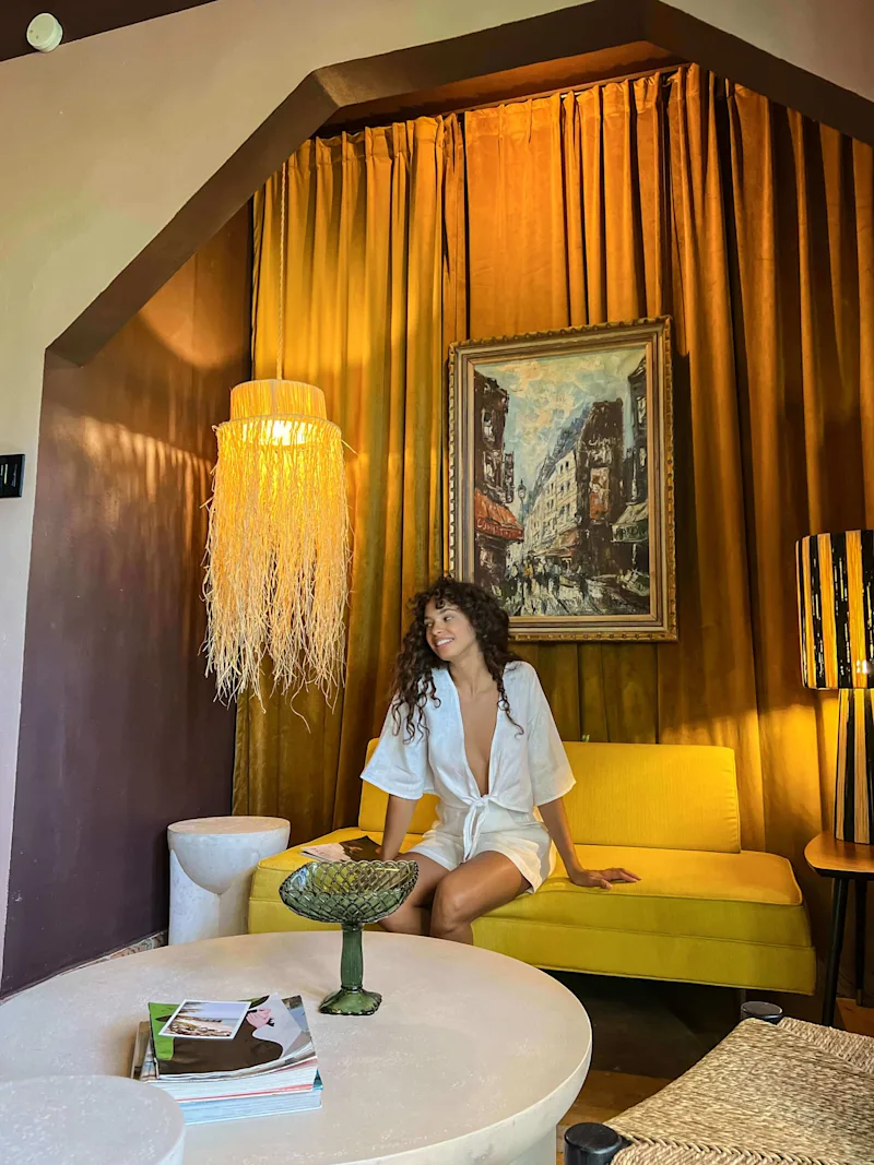 A woman smiling while sitting on a yellow sofa inside a retro-styled room featuring mustard-yellow curtains, a fringed light fixture, and framed artwork.