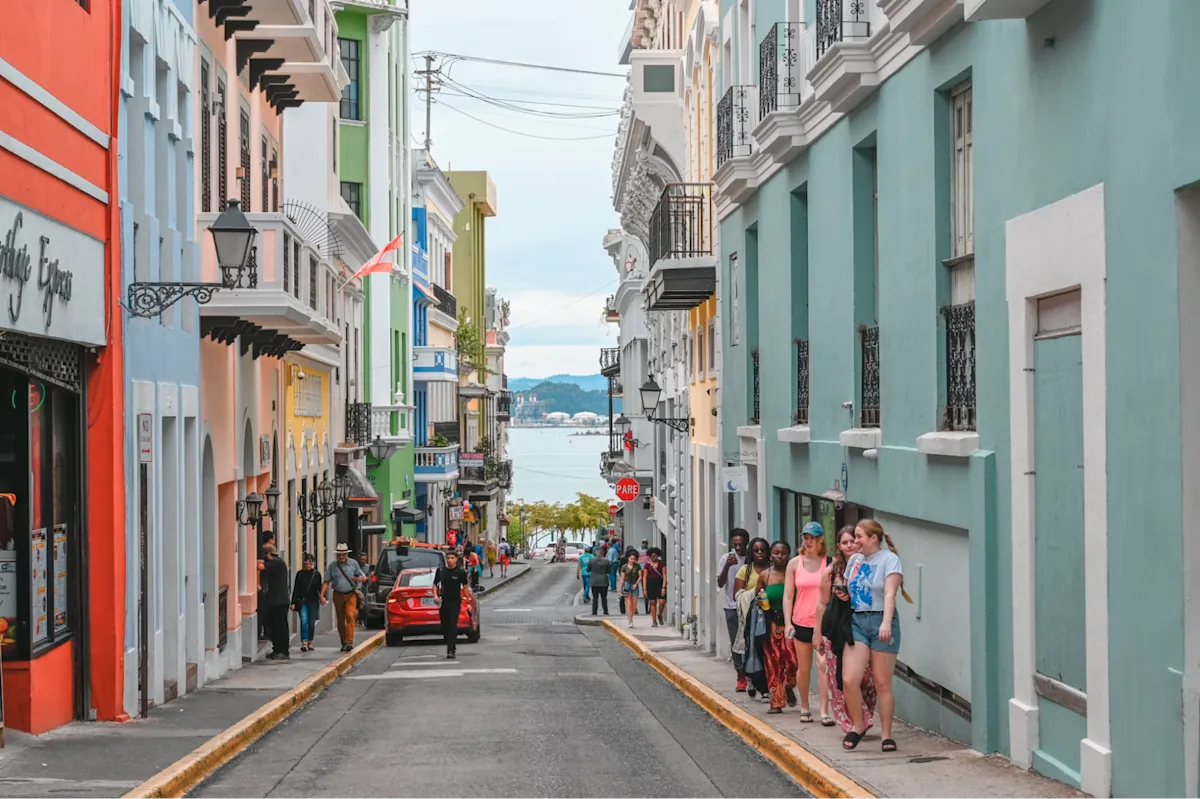 Colorful Old San Juan street with colonial buildings, balconies, pedestrians, and a view of the bay in the distance.