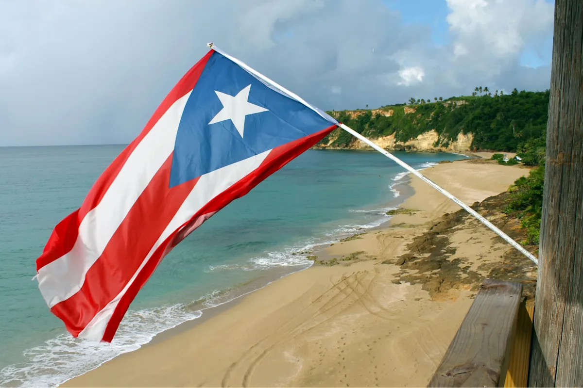 A Puerto Rican flag waves over a quiet sandy beach with turquoise water, rocky cliffs, and a cloudy blue sky in the background.