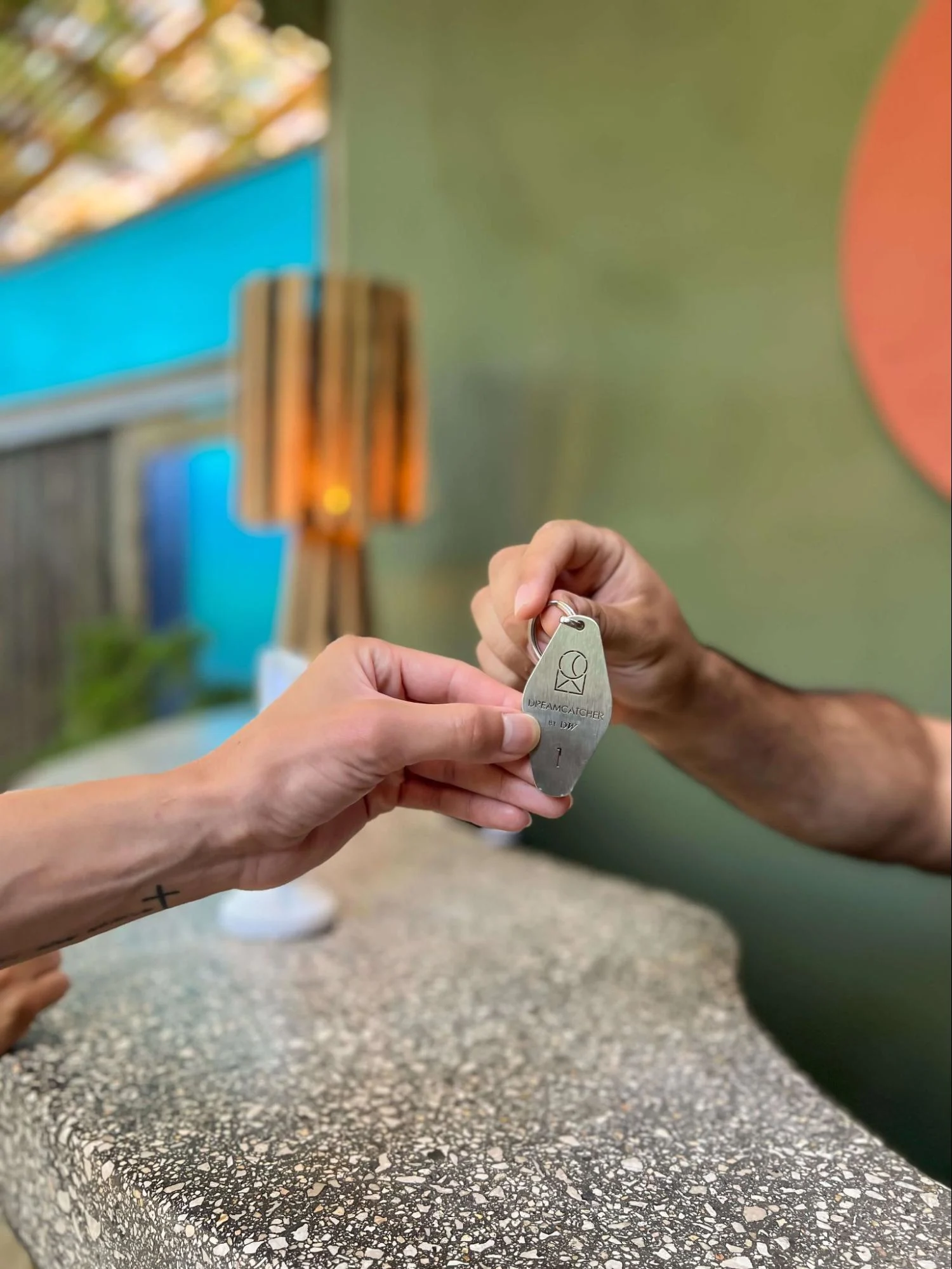 Close-up of a person handing over a metal hotel room key labeled "Dreamcatcher" at a counter.