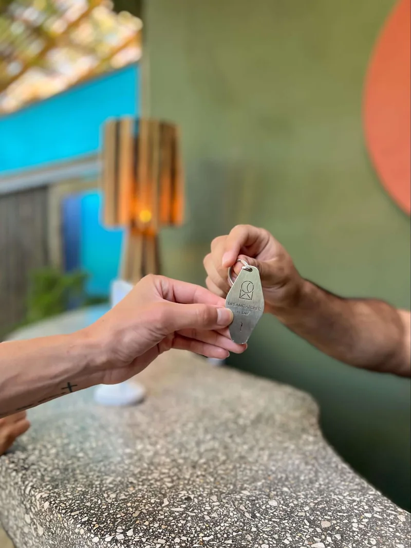 Close-up of a person handing over a metal hotel room key labeled "Dreamcatcher" at a counter.